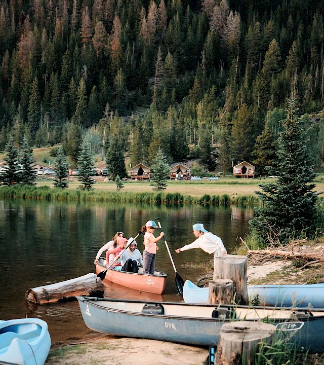 A group of people on a small orange boat near a lake, with others in blue boats, a wooden dock, and a forested hillside in the background, at a sunny day.