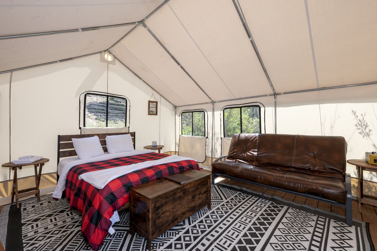 A cozy safari-style tent interior with a wooden bed, red plaid blanket, brown sofa, and large window panels.
