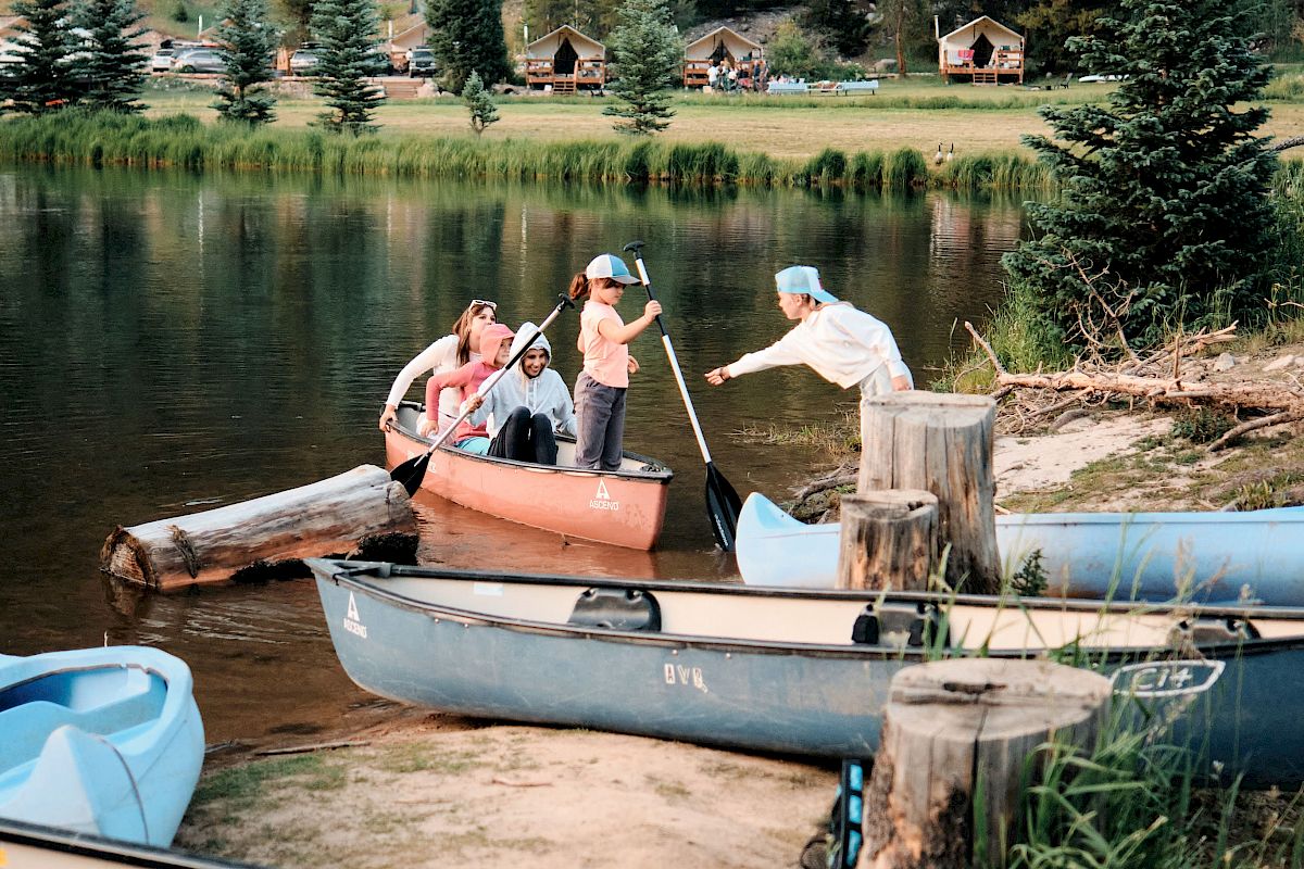 A family paddles in a canoe on a calm mountain lake, with cabins and a dense pine forest rising behind them.