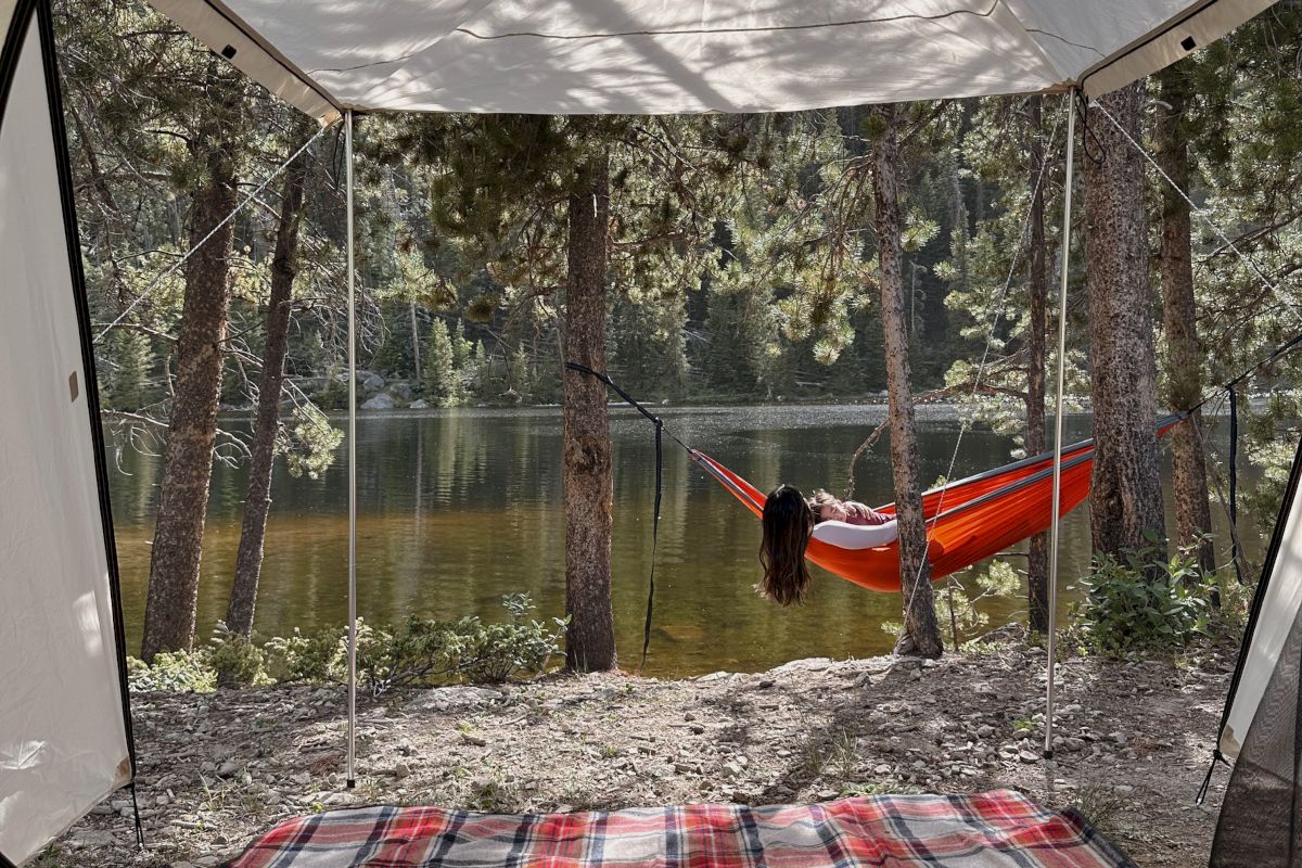 Inside an outdoor camping tent, a plaid blanket lies near the opening, facing tall trees, a lake, and a red hammock.