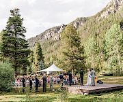 An outdoor wedding ceremony with a white tent, seated guests, a couple at a stage, and tall mountain scenery in the background.