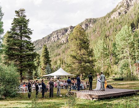 An outdoor wedding ceremony with a white tent, seated guests, a couple at a stage, and tall mountain scenery in the background.