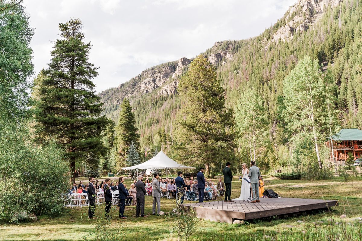 An outdoor wedding ceremony on a wooden stage with a white tent, guests, and tall mountains in the background.