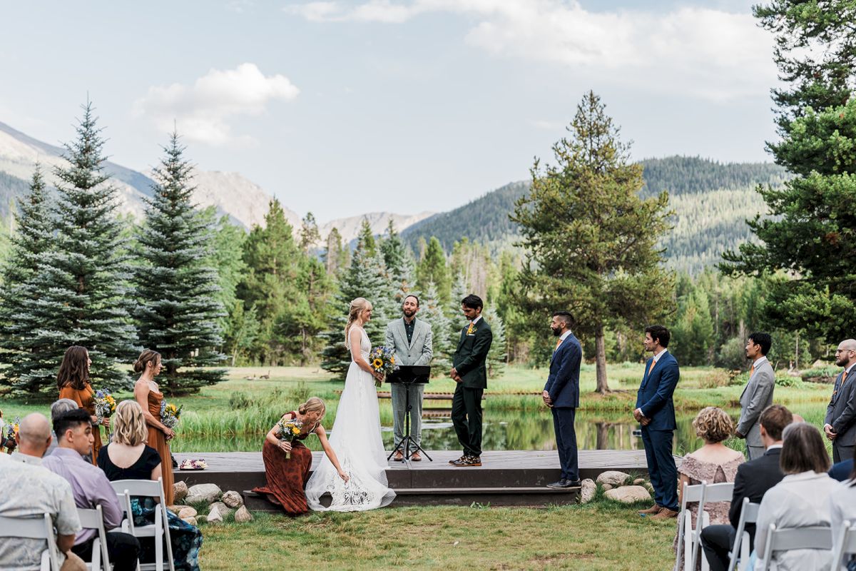 Outdoor wedding ceremony by a pond with a bride and groom at the wooden stage, surrounded by guests and mountain scenery.