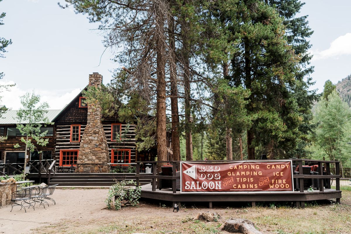 Rustic log cabin patio with banners reading &ldquo;Red Dog Saloon,&rdquo; &ldquo;Camping/Glamping,&rdquo; plus &ldquo;Ice,&rdquo; &ldquo;Cabins,&rdquo; and &ldquo;Firewood.&rdquo;
