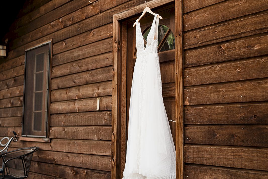 A white wedding dress hangs in an open door of a rustic wooden building, with a bouquet and shoes on the porch.