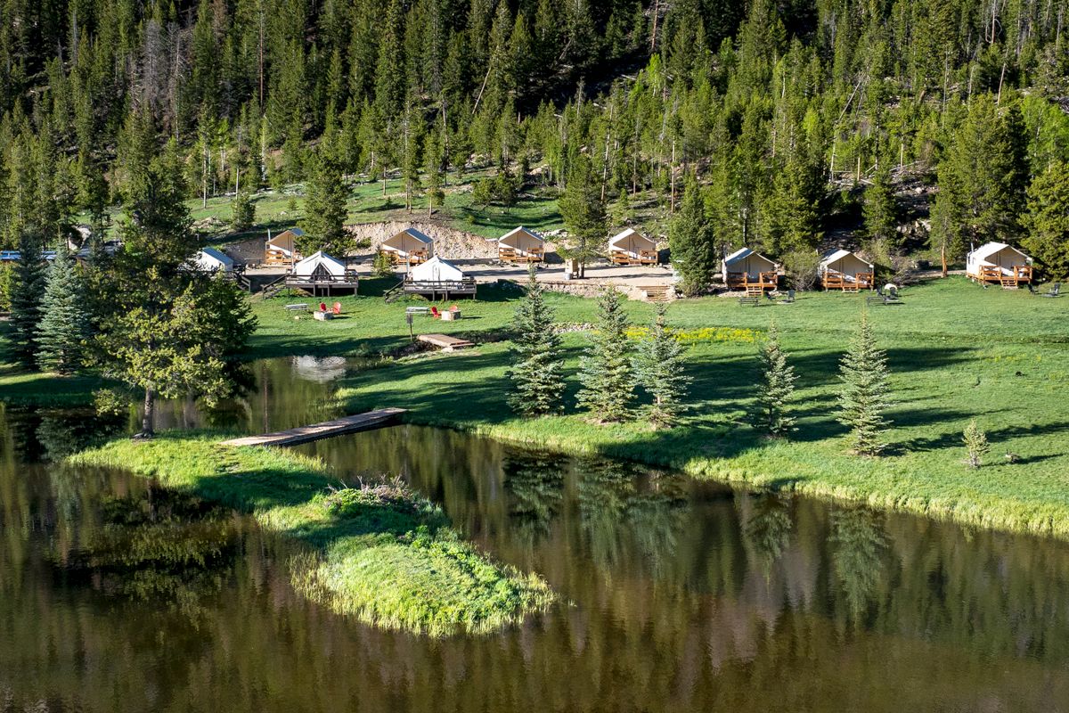 A forested alpine resort with several wooden cabins along a grassy pond, surrounded by tall evergreen trees and mountain scenery.