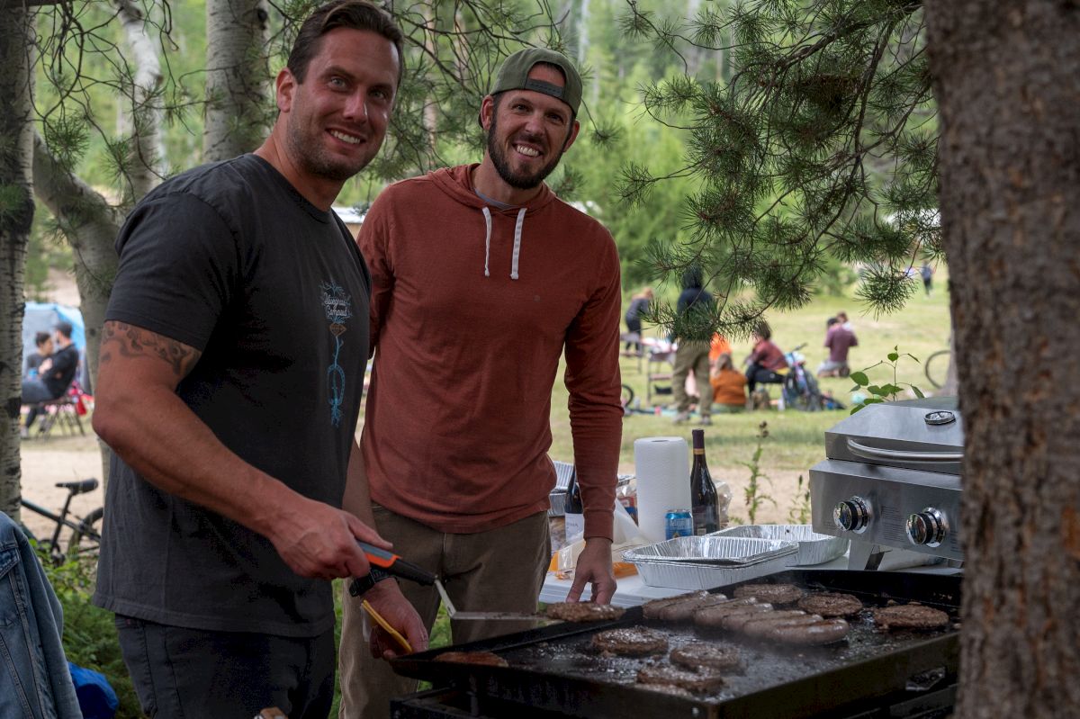 Two friends smile at an outdoor BBQ picnic in the woods, standing by a grill while others relax in the background.