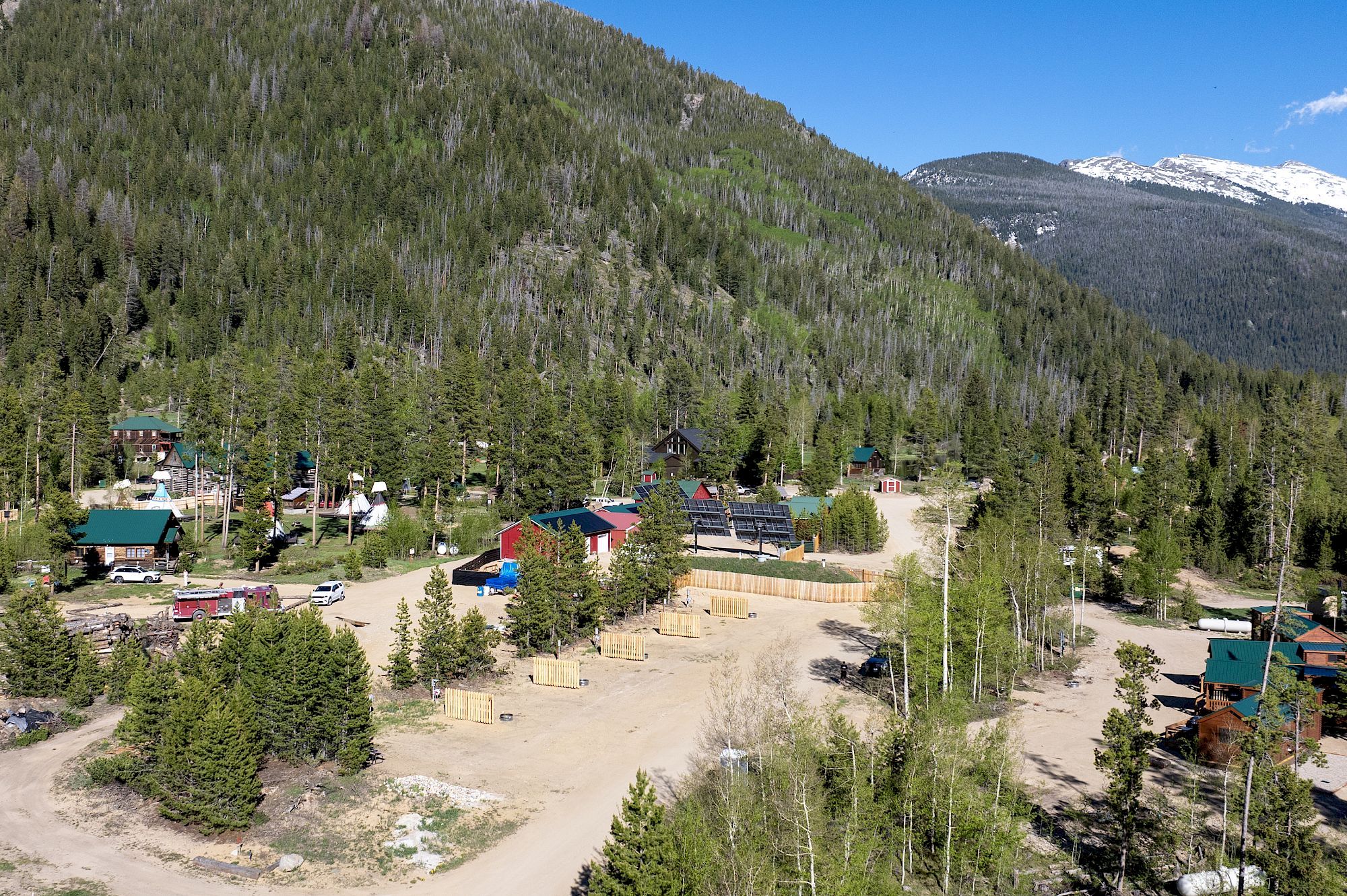 A mountain valley town nestled in dense forest, with cabins, a dirt road, parked vehicles, and snow-dusted peaks in the background.