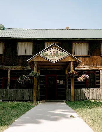 Historic wooden ranch lodge with a &ldquo;Carapaho Valley Ranch&rdquo; sign, flower planters, and wagon wheels in front.