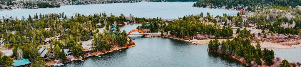 Aerial view of a calm river through a lakeside village, framed by pine forests and rugged mountains under cloudy skies.