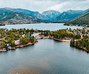 Aerial view of a calm river through a lakeside village, framed by pine forests and rugged mountains under cloudy skies.