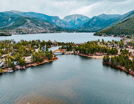 Aerial view of a calm river through a lakeside village, framed by pine forests and rugged mountains under cloudy skies.