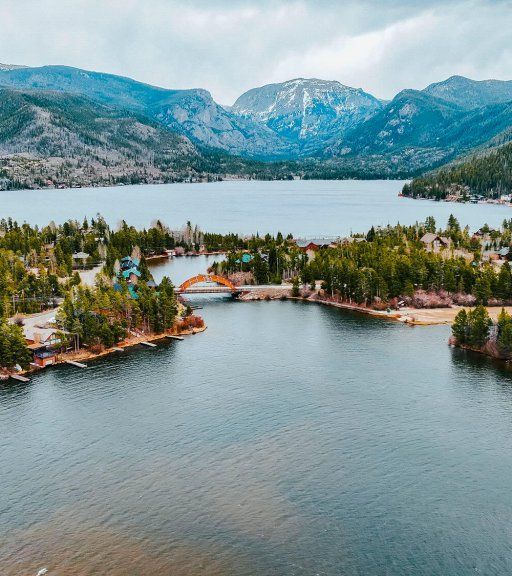 Aerial view of a calm river through a lakeside village, framed by pine forests and rugged mountains under cloudy skies.