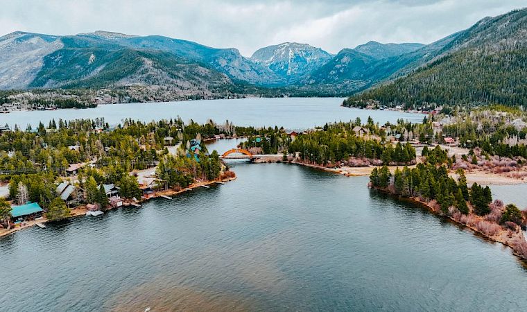 Aerial view of a calm river through a lakeside village, framed by pine forests and rugged mountains under cloudy skies.