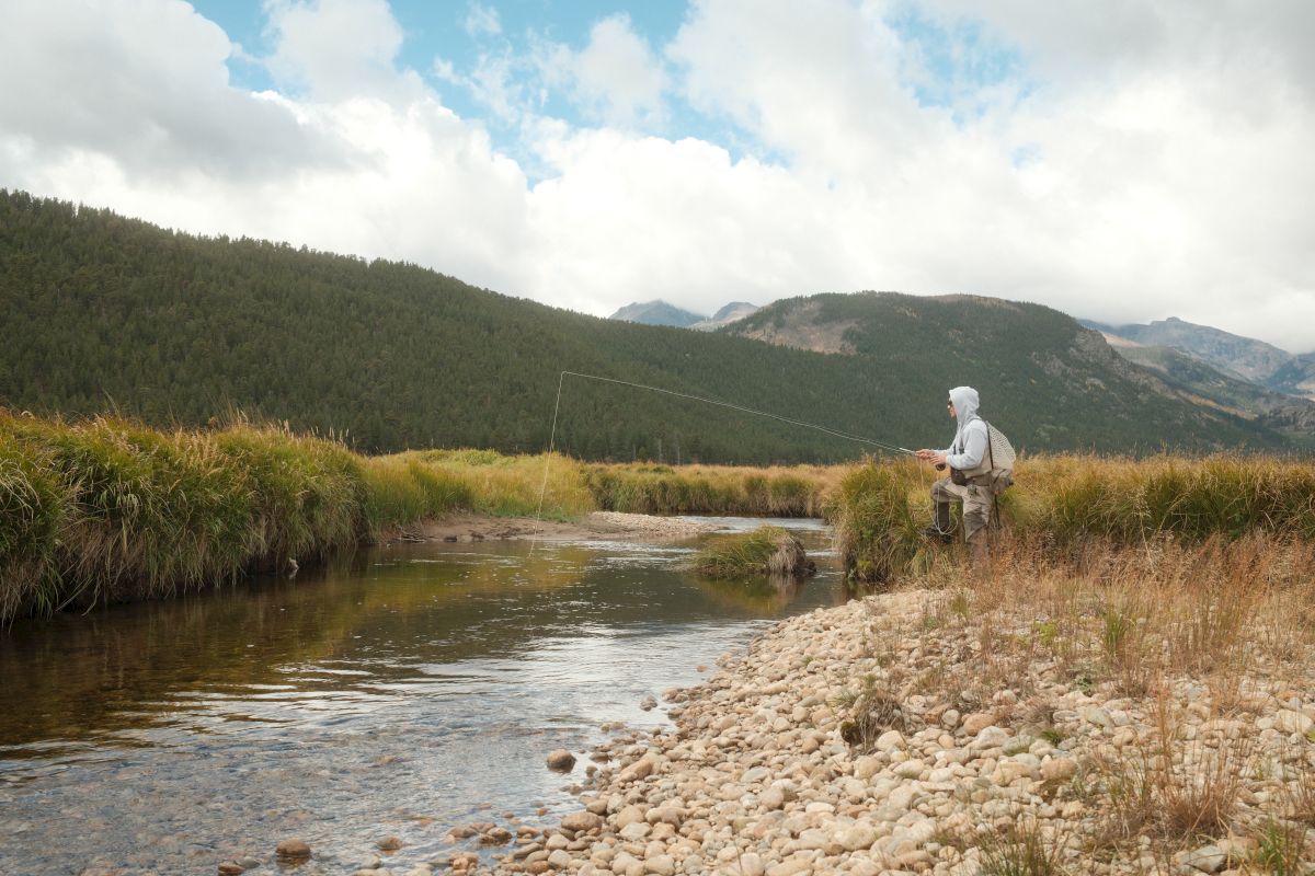 A hiker in a hoodie sits by a river, holding a fishing net, with forested mountains under a cloudy sky.