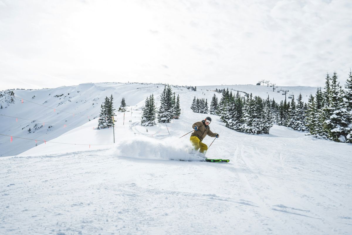 A skier carves through fresh powder on a snowy mountain slope, with lift chairs and pine trees in the background.