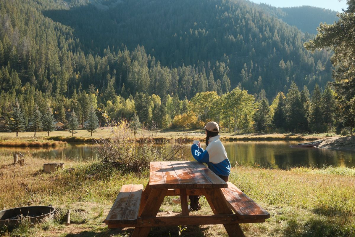 A person relaxes at a wooden picnic bench by a pond, facing misty forested mountains and golden autumn trees.