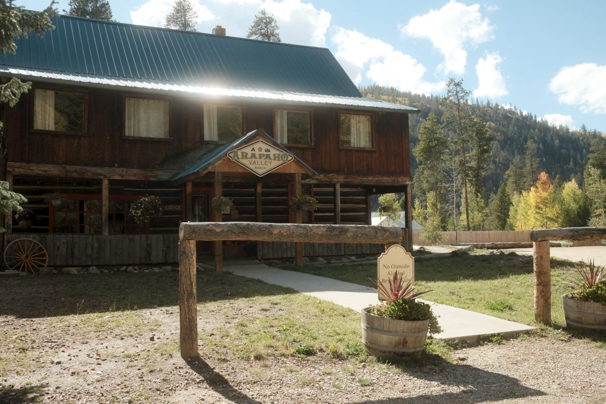 Arapaho Valley Ranch lodge with a large metal roof, wooden porch, and &ldquo;Arapaho&rdquo; sign amid forest hills.