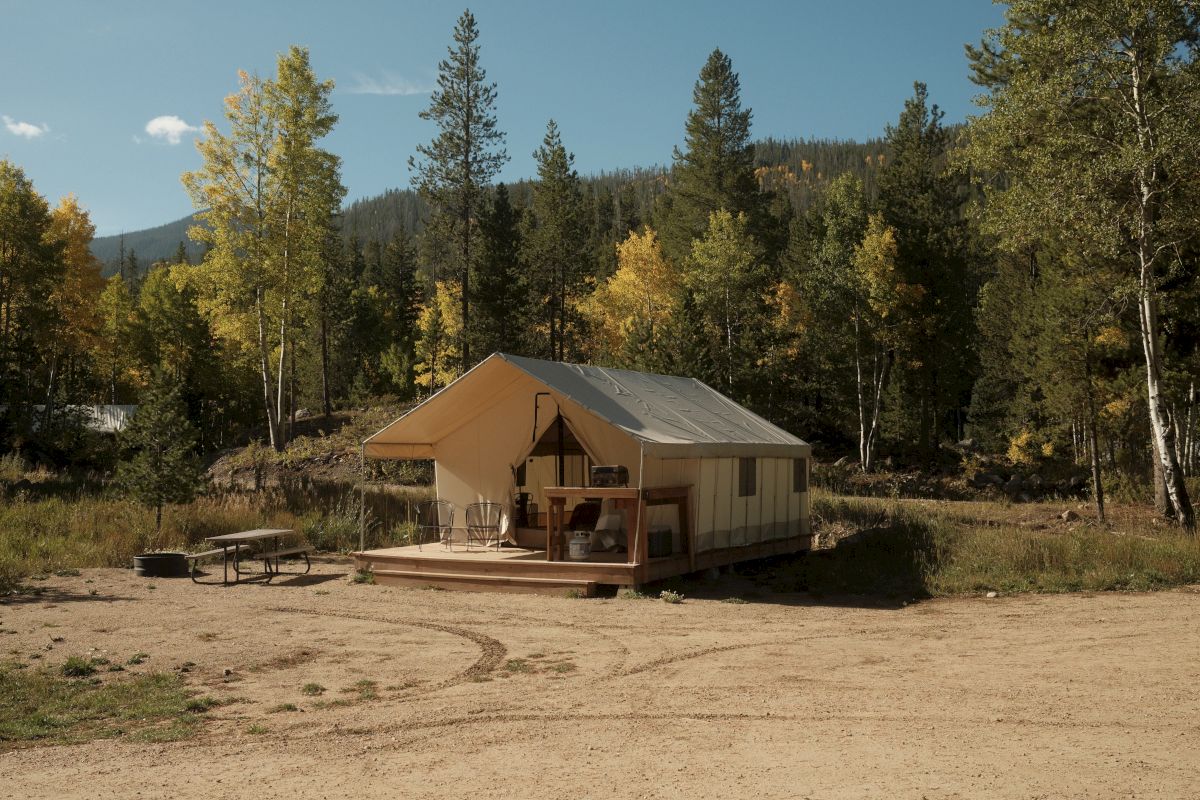 A beige canvas tent sits on a campsite in a grassy clearing, surrounded by autumn trees and a distant mountain ridge under blue sky.