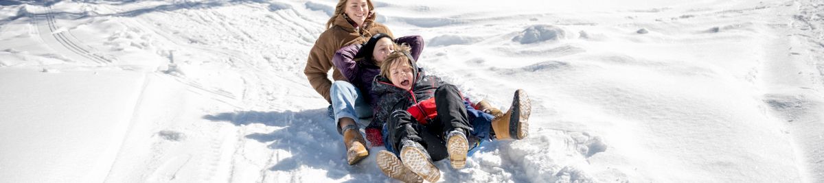 A child and two friends sled downhill on a snowy trail through a winter forest, laughing in the sunshine.