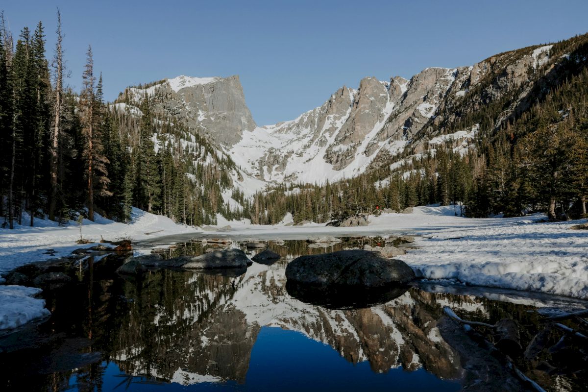 Snowy mountain valley with pine trees, rocky cliffs, and a frozen waterway reflecting the landscape in the calm pond.