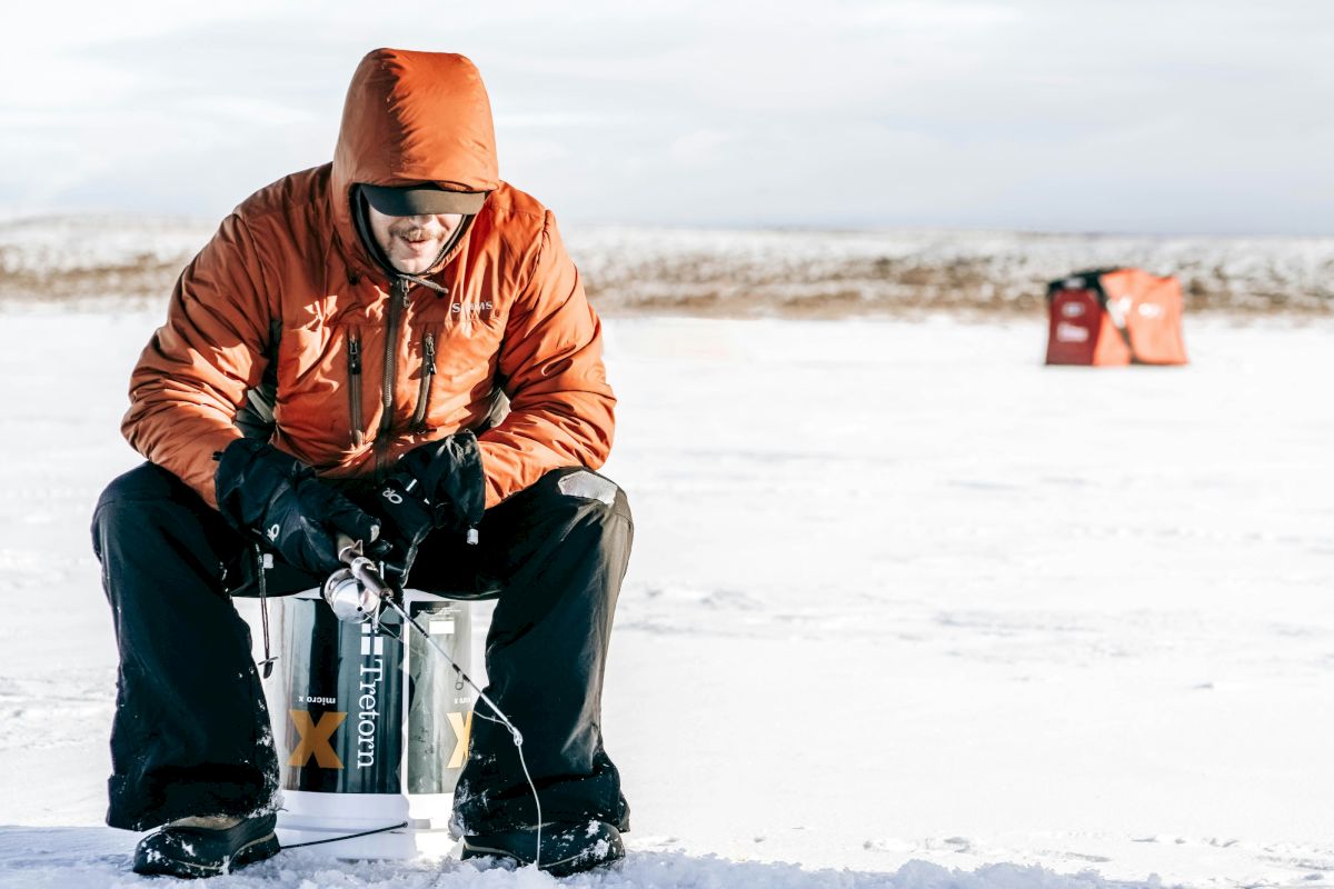 A hooded person in an orange jacket sits on a snowy beach, bundled up against the cold with gear nearby.