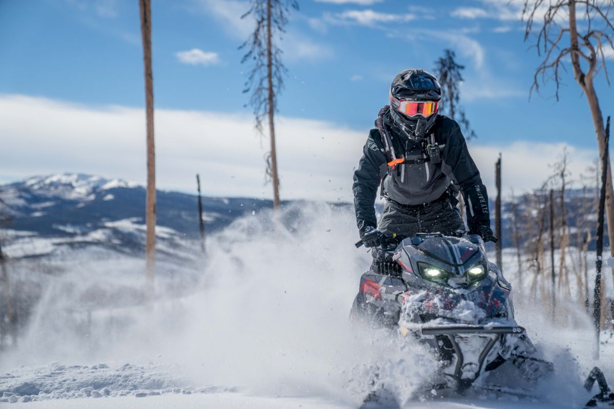 A helmeted rider drives a snow-splashing ATV through a forest trail, with mountains and blue sky in the background.