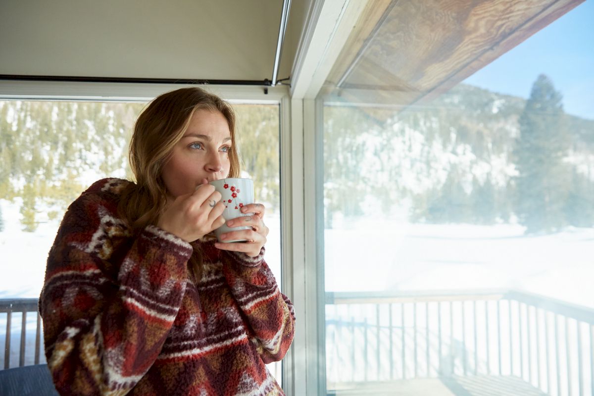 Woman in a striped sweater sips a mug by a window with snowy trees outside, enjoying a warm drink on a winter day.