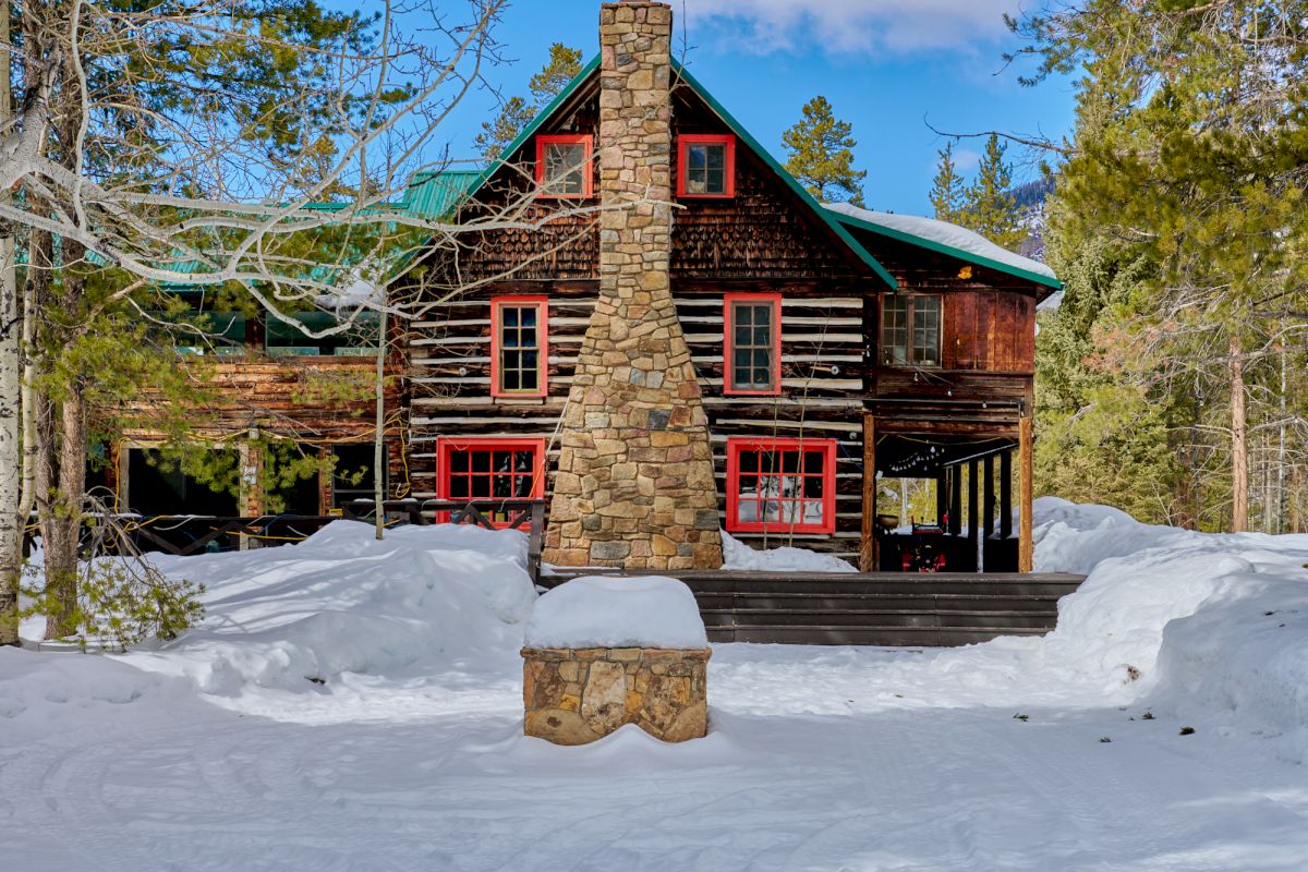A rustic log cabin with a tall stone chimney, green metal roof, and red-trim windows sits in snowy woods under blue skies.