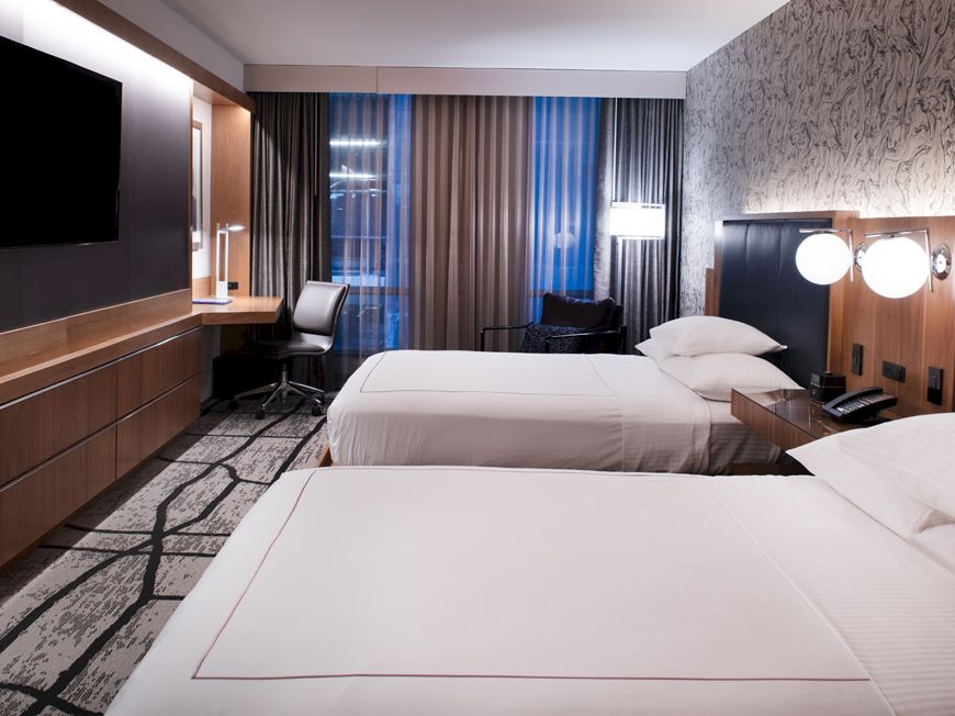 A modern hotel room with twin beds, dark wood panels, and floor-to-ceiling curtains by a city-view window.