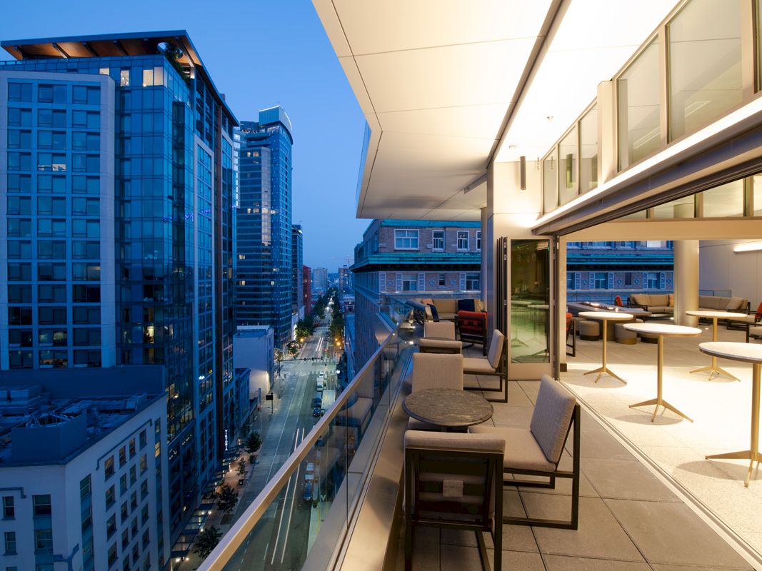 Modern rooftop terrace overlooks a downtown street, with outdoor seating and tall office buildings under a blue sunset sky.